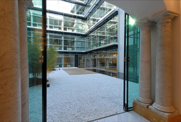 The image showcases a modern courtyard enclosed by a glass-fronted building. The perspective is from inside a classical-style room, framed by marble columns and an arched ceiling, looking out through glass doors. The courtyard features a minimalist design with a white gravel surface, a small wooden deck area, and greenery, including bamboo plants. The building’s exterior is made up of large glass panels, creating a sleek and contemporary contrast to the classical interior elements.