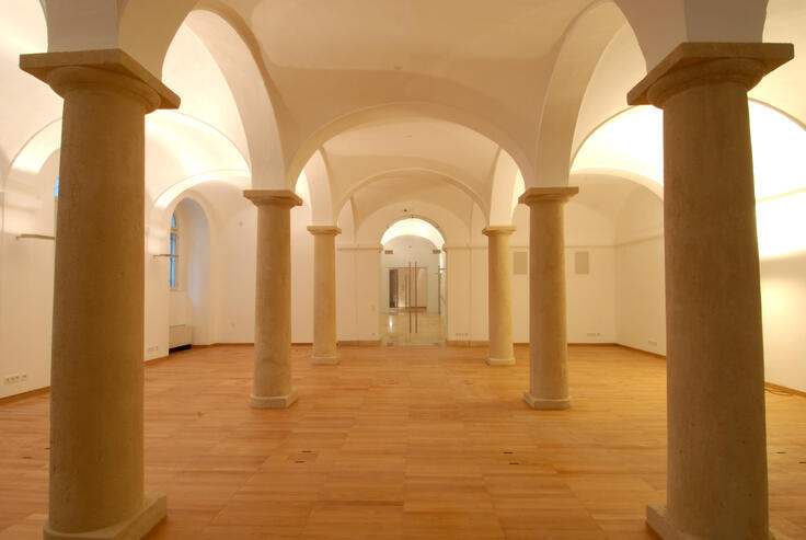 The image shows a spacious hall with classical architectural elements. The room features large, evenly spaced stone columns supporting a series of vaulted ceilings. The floor is made of warm-toned wood, contrasting with the white walls and ceilings. The lighting is soft and warm, enhancing the historical ambiance of the space.