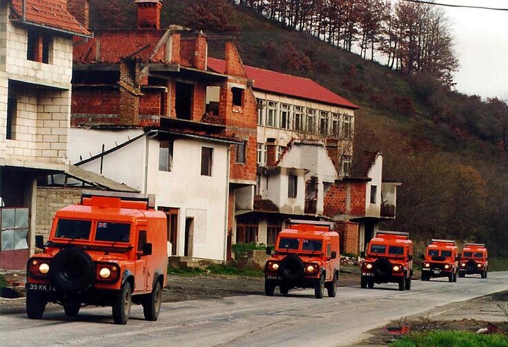 Convoy of orange armored vehicles passing through a war-torn street with damaged buildings.