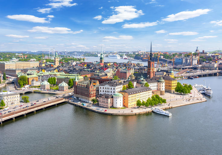 Aerial view of a vibrant cityscape by the water, with colorful buildings, a tall spire, and boats docked along the shore.