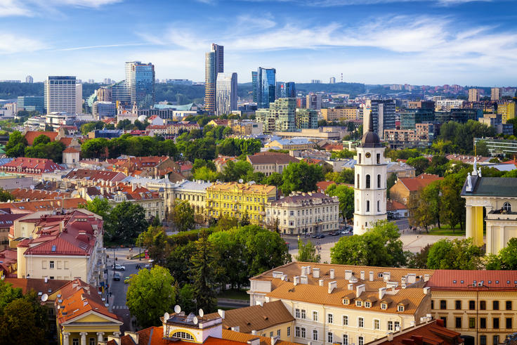 Skyline of Vilnius with colorful historic buildings, lush green trees, and modern skyscrapers under a blue sky.