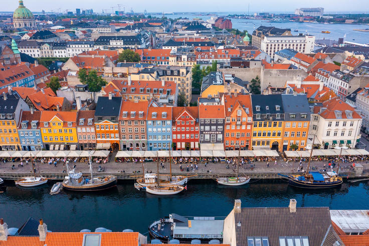 Colorful buildings line a canal in a European city, with boats moored along the water and a cityscape in the background.
