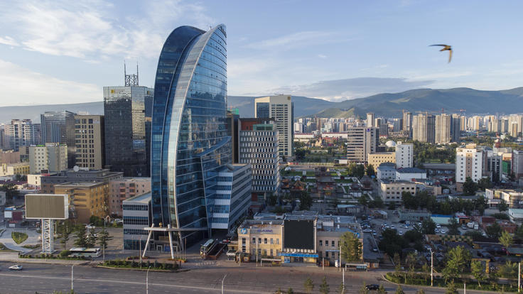 Modern cityscape with a distinctive blue glass skyscraper, surrounded by smaller buildings and mountains in the background.