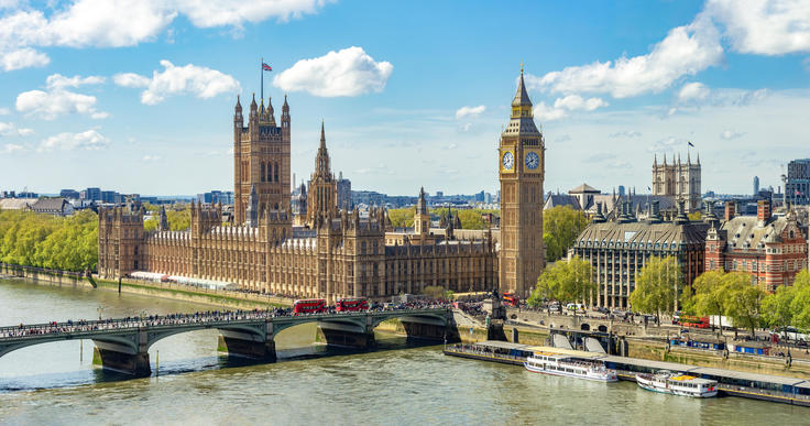 London's Houses of Parliament and Big Ben by the River Thames under a bright blue sky.