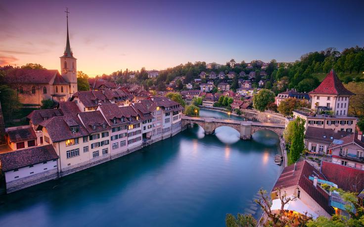 Charming town by a river at dusk, with a bridge, rooftops, and a church spire under a colorful sky.