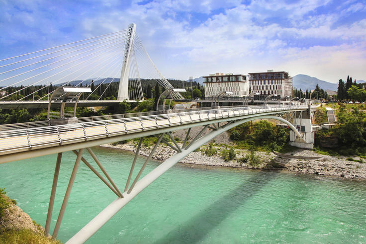Cable-stayed bridge over turquoise river with city buildings and mountains in the background.