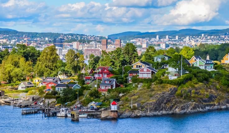 Colorful houses on a lush hillside by the sea, cityscape visible in the background, under a partly cloudy sky.