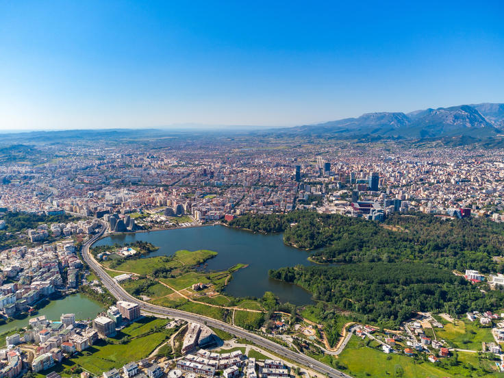 Aerial view of a city with a large green park and lake, surrounded by dense buildings, under a clear blue sky.