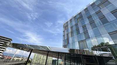 The photo shows a modern glass building reflecting a blue sky, a shiny canopy, and a train station with tracks and a red train in the background. The scene feels open and contemporary.