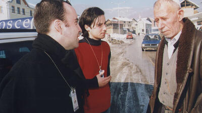 Two individuals with OSCE badges speaking to a man on a snowy street in a rural area.