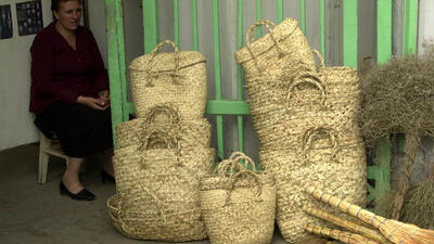 A woman sitting next to handmade woven baskets and brooms displayed for sale near a green gate.