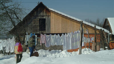 Two people stand by snowy clotheslines with laundry, in front of a rustic wooden barn.