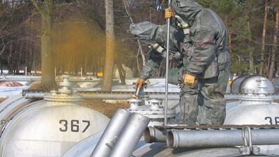 Workers in hazmat suits inspecting gas tanks with yellow fumes nearby.