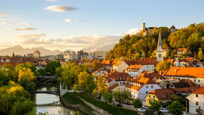 Historic city with orange rooftops, church spire, river, and lush trees under a golden evening sky.