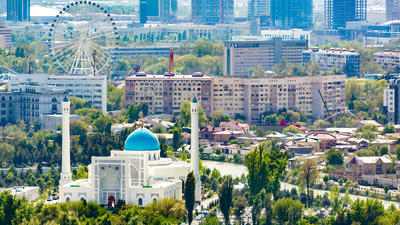 Tashkent, Uzbekistan cityscape with a mosque, characterized by a blue dome and two minarets, surrounded by trees and urban buildings. Ferris wheel in background.