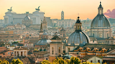 Skyline of Rome at sunset, featuring historic domes and buildings under a warm, orange sky.