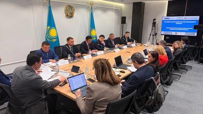 Meeting with people around a long table, two Kazakhstan flags behind, presentation screen on the side.