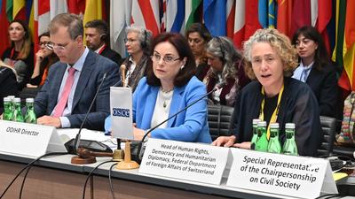 Three people sit at a conference table with microphones in front of national flags.
