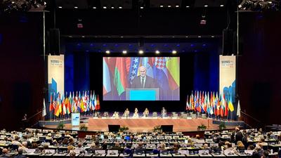 A large conference hall with a speaker at a podium, international flags, and attendees seated.