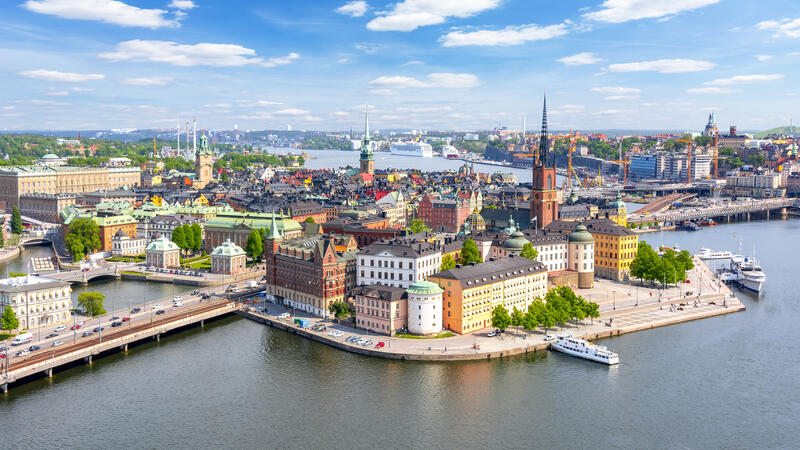 Aerial view of a vibrant cityscape by the water, with colorful buildings, a tall spire, and boats docked along the shore.