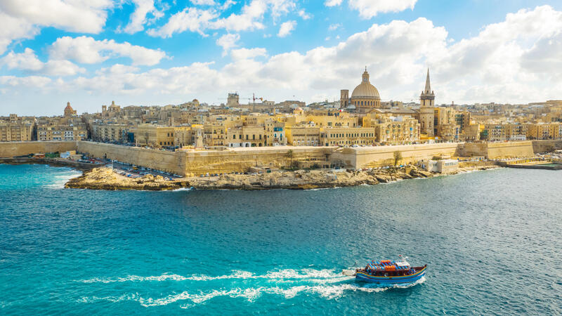 Valletta's skyline with historic buildings under a blue sky, viewed from the sea with a small boat in the foreground.