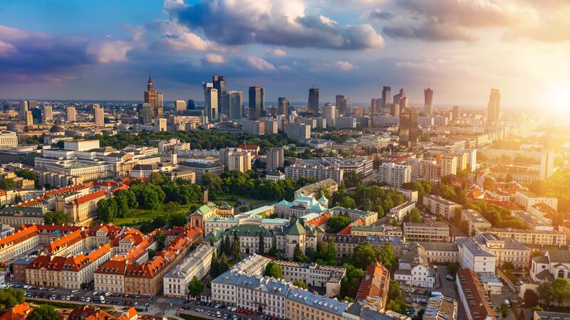 Aerial view of a cityscape at sunset, with skyscrapers, green parks, and historic buildings beneath a cloudy sky.
