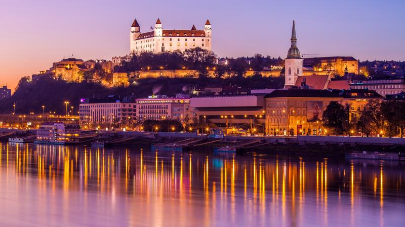 Bratislava Castle illuminated at sunset, reflecting on the Danube River, with a clear purple sky.