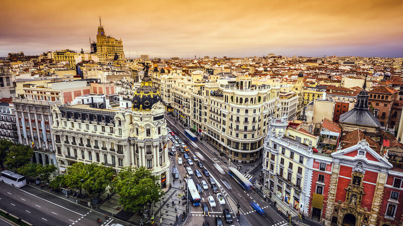 Bustling cityscape with ornate buildings under an orange sunset sky, busy street with cars and buses.