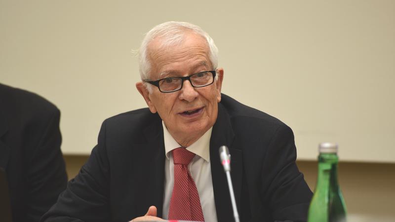 A man in a suit and tie speaks at a conference table, a microphone in front of him.