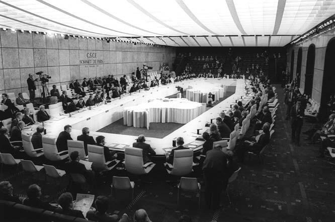 A black-and-white photograph of the CSCE Paris Summit, showing a large conference room with delegates seated around a U-shaped table arrangement. The backdrop includes the text 'CSCE Sommet de Paris,' and the space is filled with officials, media personnel, and observers.